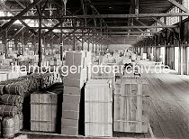 586_662a Die Waren stehen gut und sicher verpackt in einem Lagerschuppen des Hamburger Hafens - im Hintergrund transportieren Hafenarbeiter Kisten mit Elektrokarren. Sackkarren mit den gebogenen Handgriffen sind in den Gngen des Lagers abgestellt.