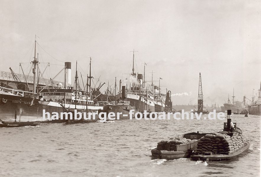 0953981historisches Foto vom Hamburger Hafen - Blick in den Hansahafen; links liegen Frachter am Oswaldkai - eines der Schiffe wird ber einen schwimmenden Getreideheber entladen, daneben ist ein grosser Schwimmkran zu erkennen. Dahinter ist als Silhouette die Wohnblocks auf der Veddel zu erahnen. Im Vordergrund ein mit Scken beladener Schlepper, der eine Schute im Schlepp hat.
