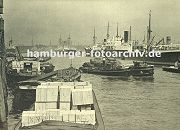 0953983 Im Hansahafen werden Schuten beladen - hoch getrmt liegen Holzkisten in einem Kahn; ein anderes Transportschiff wird mit seiner Ladung von einem Schlepper zu seinem Bestimmungsort gebracht. Im Hintergrund Kirchtrme der Hansestadt Hamburg.