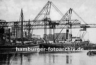 k_0954022 Frachtschiffe liegen im Harburger Seehafen am Hafenkai - die Kranausleger sind heruntergelassen und beladen oder entladen die Schiffe. Eine Schute hat am Dampfschiff lngsseits fest gemacht und wird mit dessen bordeigenem Ladebaum beladen; im Hintergrund Harburger Fabrikgebude.