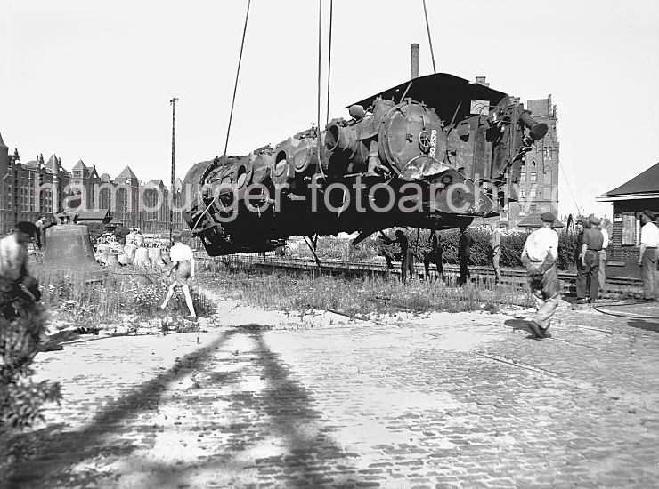 Kriegsschden - Beseitigung - Lokomotive Sandtorhafen, Speicherstadt 1948  441_03 Eine im Krieg zerstrte Lokomotive wird 1948 von einem Kran im Sandtorhafen geborgen; im Hintergrund die Gebude der Speicherstadt am Brooktorkai - rechts ist ein Ausschnitt des Kaispeichers B zu erkennen. Die Kirchenglocken im linken Bildrand wurden whrend der Kriegszeit auf Befehl in den Kirchentrmen abgehngt und teilweise auch im Freihafen zwischengelagert - sie sollten eingeschmolzen werden.