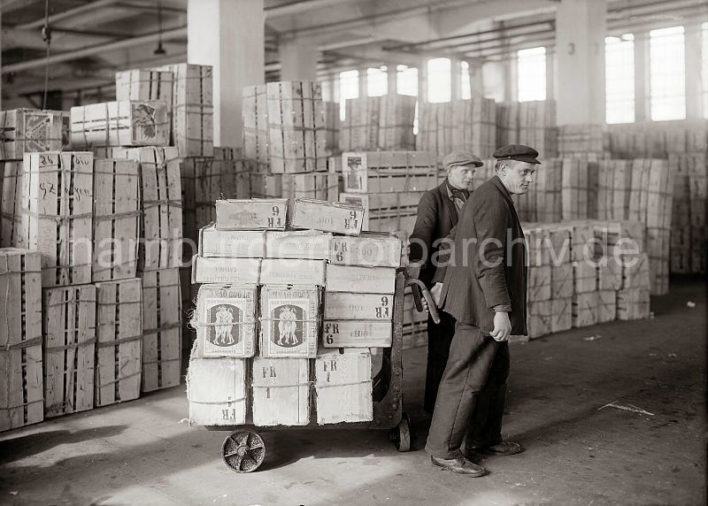 460_419 Zwei Lagerarbeiter transportieren Apfelsinenkisten mit einer Karre im Lager des Fruchtschuppens im Hamburger Hafen. Im Hintergrund sind die Kisten mit den S�dfr�chten gestapelt.  Historische Hamburgbilder + Hamburgensien auf Fotoleinwand und Posterprint; als Geschenk fuer Buero und Wohnung.