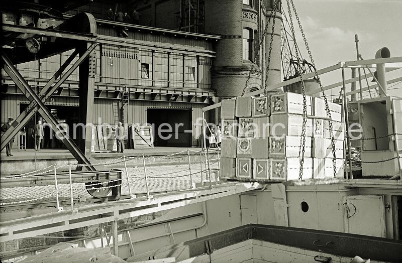 Historische Hamburgbilder + Hamburgensien auf Fotoleinwand und Posterprint; als Geschenk fuer Buero und Wohnung. Ladung l�schen eines Frachtschiffs - Apfelsinenkisten am HafenKran. 461_OF_46A Eine Hieve Apfelsinenkisten h�ngt am Ladegeschirr �ber dem Laderaum des Schiffs - auf dem Kai das Laufrad und die Schiene des fahrbaren Halbportalkrans vor dem Fruchtschuppen. Auf der Laderampe stehen die Hafenarbeiter und warten darauf, die Ladung in Empfang zu nehmen.