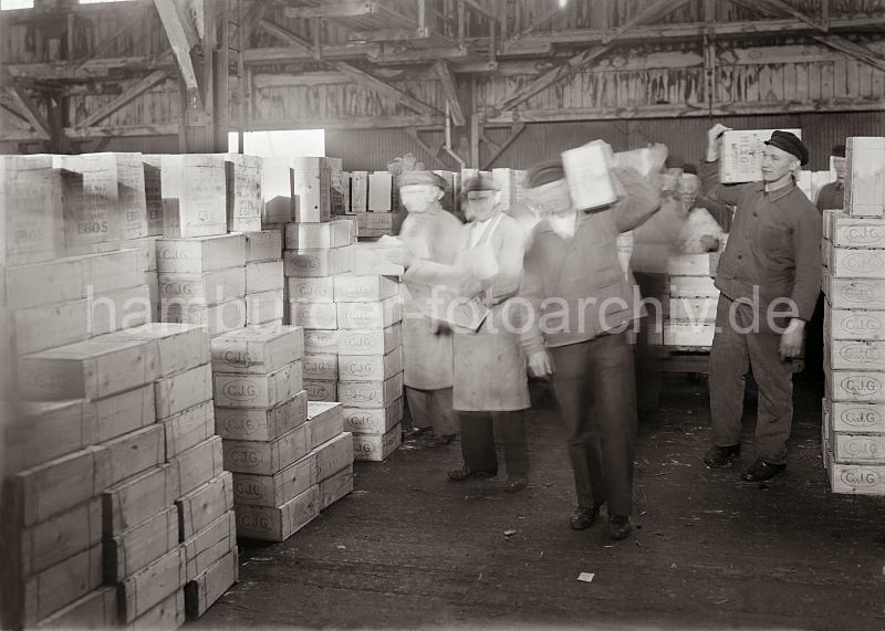 historische Arbeitsbilder aus dem Hamburger Hafen - Kisten stapeln im Fruchtschuppen; ca. 1932. Bilder von der Geschichte Hamburgs - Arbeit im Hafen Hamburg.  471_577 Die gel�schten Rosinenkisten sind am Hafenkai auf einen Transportwagen geladen worden - im Fruchtschuppen werden die Kisten von Hand abgeladen und von den Lagerarbeitern auf der Schulter zu dem Lagerplatz gebracht.