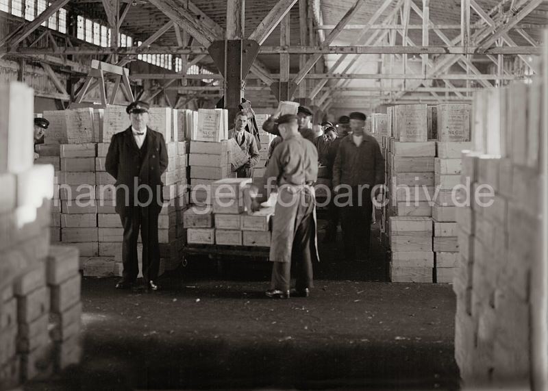historische Fotografien aus der Arbeitswelt im Hamburger Hafen, Arbeit im Fruchtschuppen; ca. 1932. Bilder von der Geschichte Hamburgs - Arbeit im Hafen Hamburg. Alte Hafenbilder aus dem Archiv der Hamburger Hafen und Logistik AG 472_577a Die Lagerarbeiter nehmen die Kisten mit den Rosinen von dem Transportkarren auf und bringen sie auf der Schulter zur dem Lagerplatz. Die Kisten gef�llt mit getrockneten Weinbeeren sind in dem Fruchtschuppen mannshoch aufgestapelt.