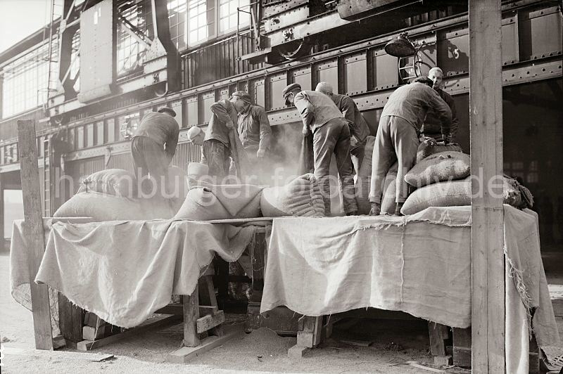 Eine Ladung Scke wird von Lagerarbeitern vor dem Lagerschuppen geleert - Laufschiene Halbportalkrne; ca. 1934 - Alte Hafenbilder aus dem Hamburger Hafen;  481_B_331 Vor dem Tor des Lagerschuppens stehen die Lagerarbeiterarbeiter auf einem Podest und leeren die Scke, die vom Frachter gelscht wurden. An der Wand ist die Laufschiene der Halbportalkrne zu erkennen, die den Hafenkai berspannen.