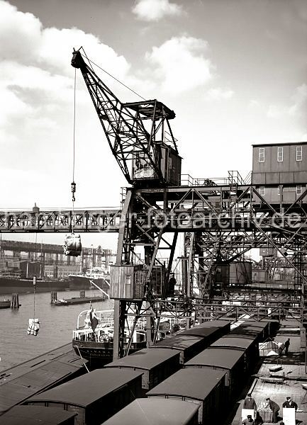 Alte Bilder aus dem historischen Hafen Hamburgs; Dreifachkran beim Lschen eines Frachters - Gterzuege am Kai. Fotos von der Geschichte des Hamburger Hafens. 483_76 Mit einem Dreifachkran wird die Ladung eines Frachters im Hamburger Hafen gelscht. Zwei Hieven Scke und eine Ladung Kisten bringt die Krananlage an Land. Unter dem Portal des Hafenkrans stehen zwei Gterzge mit geschlossenen Waggons. Auf der Rampe vor dem Lagerschuppen transportieren Hafenarbeiter Kisten mit der Sackkarre.