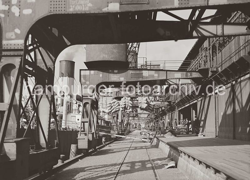 . Alte Arbeitsbilder aus dem Archiv der Hamburger Hafen und Logistik AG. Hafenarbeiter auf der Laderampe. Fotos von der Geschichte des Hamburger Hafens. 484_648a Am Kai hat ein Frachtschiff fest gemacht, aus seinem Schornstein steigt leichter Qualm. Drei Hieven Scke hngen an den Kranhaken und werden auf die Laderampe den Hafenschuppens gebracht. Dort warten die Hafenarbeiter darauf, die Ladung mit Karren in den Lagerschuppen zu bringen.