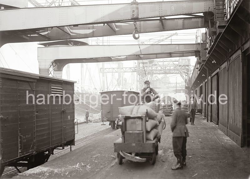 Bilder der Geschichte des Hafen Hamburgs - Fotoarchiv der Hamburger Hafen und Logistik AG.  486_585 Der Hafenkran hat gerade eine Hieve Scke auf dem Anhnger eines Elektrokarrens abgesetzt, die Ladung wird von Arbeitern vom Kranhaken gelst. Auf dem Gleis vor dem Lagerschuppen stehen geschlossene Gterwaggons.