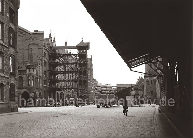 Backsteinarchitektur der Lagerhuser in der Hamburger Speicherstadt;   502_565 Blick vom Bereich des Speicherblock E und der Zollabfertigung am Kai des Zollkanals vom Brook zum Speicherblock D. Der Fahrer eines mit Scken hoch beladenen Lastwagens wird von seinem Beifahrer ber die Strasse dirigiert. Im Vordergrund fhrt ein Radfahrer ber das Kopfsteinpflaster. Rechts hinter dem Schuppendach ein Ausschnitt des Brckenportals der Brooksbrcke, die ber den Zollkanal zur Mattentwiete fhrt.