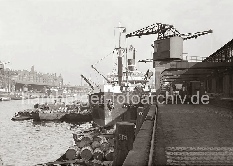 Alte Bilder aus dem Hafen Hamburgs - Fotoarchiv der Hamburger Hafen und Logistik AG (HHLA)- Halbportalkrne im Sandtorhafen / Kaiserkai - Schuten mit Tonnen beladen; ca. 1932. 510_723 Die Ladung Fsser eines Frachtschiffs am Kaiserkai des Sandtorhafens wird gelscht - mit den Halbportalkrnen werden die Holztonnen an Land gebracht - sie liegen auf der Laderampe und sind auf einem Gterwaggon gestapelt. Auf der Wasserseite des Schiffs werden Schuten mit dem bordeigenem Ladegeschirr des Frachtschiffs beladen. Im Vordergrund an der Kaimauer des Kaiserkais eine mit Fssern hochbeladene Schute. Auf der anderen Seite des Sandtorhafens die einfachen Ladekrane am Sandtorkai und die Dcher mit den Giebeln der Speicherstadt.