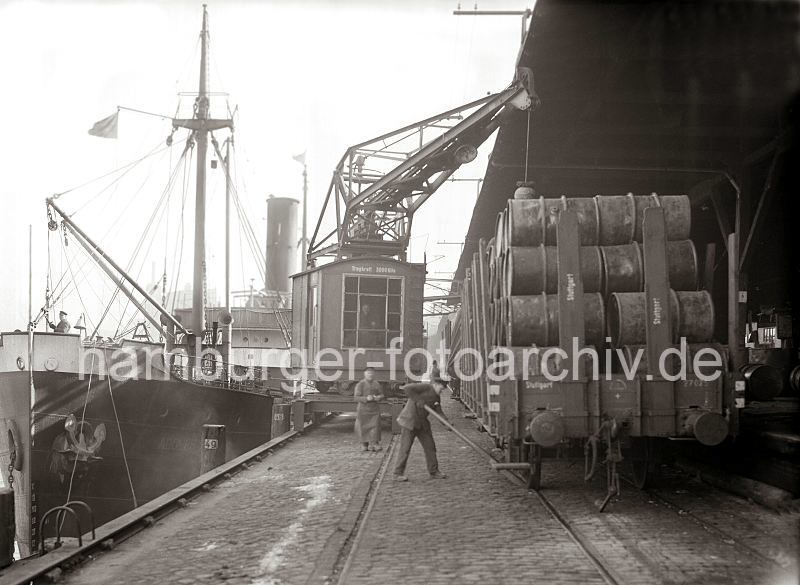 Alte Hafenmotive aus Hamburg - Fotoarchiv der Hamburger Hafen und Logistik AG (HHLA. Hafenkran am Kai - Frachter und mit Fssern beladener Gterzug; ca. 1930.  512_428 Ein auf Schienen laufender Hafenkran ldt Fssern auf einen Gterzug, der an der berdachten Rampe des Kaischuppens steht.