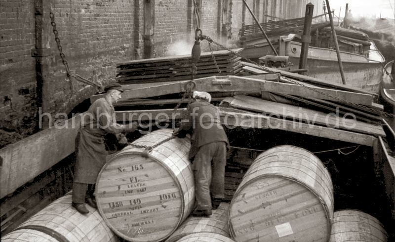Alte Hamburgfotografien aus dem Hafen und der Speicherstadt der HHLA Tabakfässer im Saalehafen - Lagerhaus G, Arbeiter legen das Ladegeschirr an; 1948.  516_0001 Die grossen Holzfässer mit Tabak sind im Laderaum der Schute gestapelt. Zwei Arbeiter legen das Ladegeschirr an dem Fass an, damit es in den Laderaum des Lagerhaus G am Saalehafen gehoben werden kann. Im Hintergrund liegen weitere Schuten an der Mauer des Ziegelgebäudes.