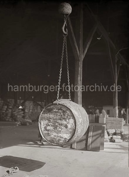 Altes Fotomaterial vom Hamburger Hafen / Archiv HHLA Gterumschlag - Verladung von Fssern, Ladegeschirr; ca. 1930. 525_438 Ein Fass hngt an der Kette des Ladegeschirrs im Haken des Krans. Im Hintergrund lagern Kisten und Scke.