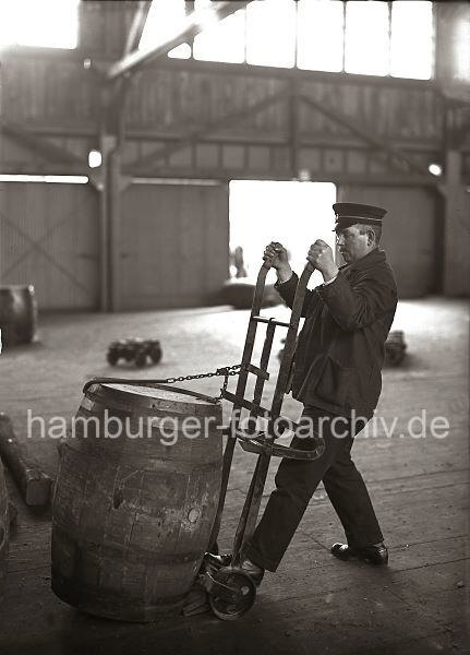 Altes  Bildmaterial vom Hamburger Hafen / Archiv HHLA Transport von einem Fa, Selbstladekarre; ca. 1930. 527_69 Bei einer Selbstladekarre wird das Fass beim Beladen mit einem eisernen Haken, der an einer Kette befestigt ist, auf die Handkarre gezogen. So kann der Lagerarbeiter die Tonne ohne grossen Kraftaufwand an ihren Bestimmungsort transportieren.