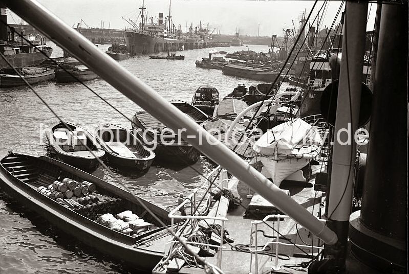 Altes Fotodokument vom Hamburger Hafen - Be- und entladen von Frachtschiffen im Hafen Hamburg - Schuten mit Ladung, Kisten, Scke Fsser; ca. 1932. 528_226 Frachter haben am Hafenkai fest gemacht, sie werden ber lngsseits liegende Schuten be- und entladen. Fsser, Scke und Kisten liegen in dem offenen Laderaum der Khne, die mit Hilfe eines Schleppers durch den Hafen gezogen werden. Weitere Frachtschiffe liegen an den Holzdalben in der Mitte des Hafenbeckens.