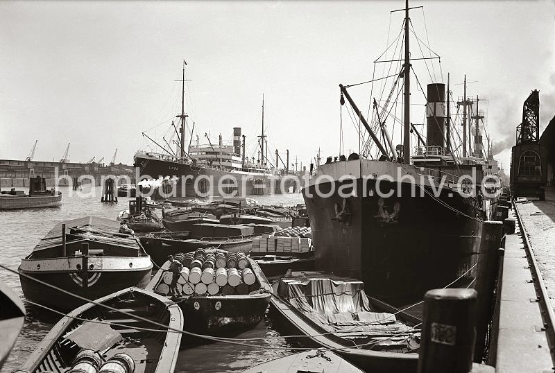 otos von der Hamburger Hafengeschichte / Bildarchiv der HHLA Gterumschlag im Hafen Hamburg - Schuten beladen mit Ladung, Kisten und Fssern; ca. 1932.  529_227 Frachter liegen am Hafenkai - Holzstmme sind zum Schutz der Kaimauer und der Schiffe in den Hafengrund gerammt; sie sind nummeriert - so kann der Liegeplatz des Frachtschiffs bestimmt werden. Hafenkrane laufen auf Schienen am Rand des Kais. ber bordeigenes Ladegeschirr lschen die Frachter ihre Ladung auf lngsseits liegende Schuten oder nehmen neue Ladung auf. Die Lastkhne sind mit Fssern und Kisten hoch beladen; ein Schlepper liegt unter Dampf zwischen den Khnen, die keinen eigenen Antrieb haben und gezogen werden mssen.