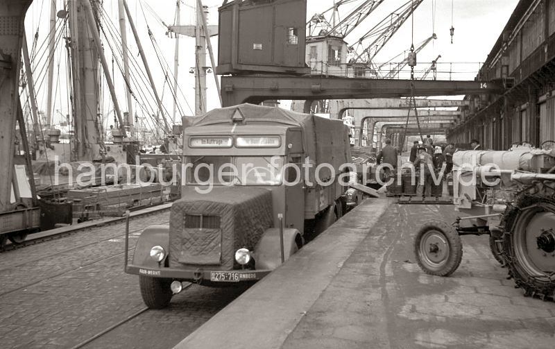 Bilder der Hamburger Hafengeschichte Lastwagen mit Plane an der Laderampe - Korbflaschen und Traktor auf der Laderampe im Rohafen; ca. 1952. 530_0007 Ein LKW der FAUN Werke Nrnberg steht an der Laderampe des Rohafens, die Plane der Ladeflche des Lastkraftwagens ist geschlossen - eine Anzeige ber der Fahrerkabine weist darauf hin das der Laster im Auftrag der Bundesbahn fhrt. Ein Traktor steht auf der Rampe bereit zur Verladung - sein Motor ist mit dicker Pappe fr den Transport geschtzt.  Im Hintergrund verladen Hafenarbeiter grosse Korbflaschen auf eine Stauerplatte, die mit dem Halbportalkran an Bord des Schiffes gehievt wird.