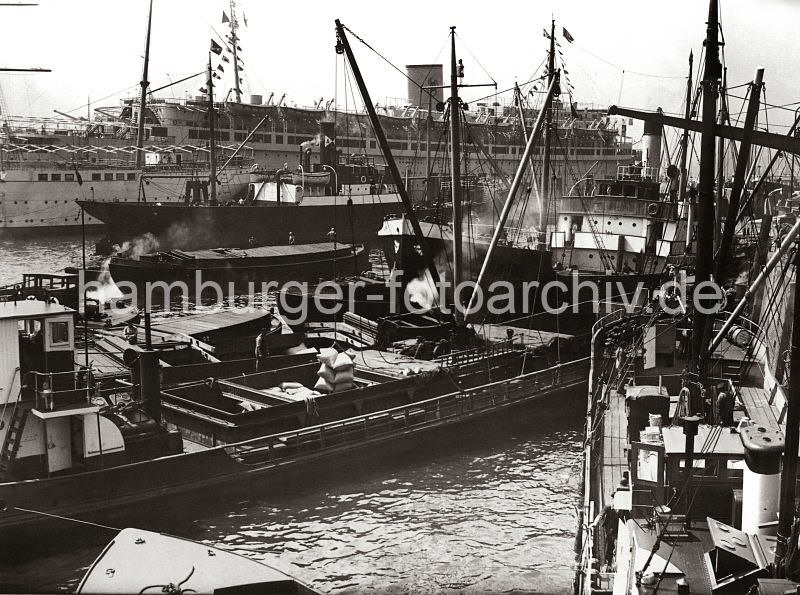 Hamburgensien aus dem Hamburger Hafen - Motorschiffe im Hafen Hamburgs - Frachter und Schuten am Johannisbollwerk / Vorsetzen - Passagierschiff Wilhelm Gustloff.  537_061_89 Motorschiffe haben vor dem Kai an den Vorsetzen fest gemacht - die Ladung wird auf Binnenschiffe, die lngsseits liegen gelscht oder von den Lastkhnen aufgenommen. Eine Hieve Scke hngt im Ladegeschirr des Ladebaums und schwebt ber dem Laderaum des kleinen Frachters.  Im Hintergrund das 1938 auf der Hamburger Werft Blohm & Voss gebaute NS-Passagierschiff Wilhelm Gustloff.