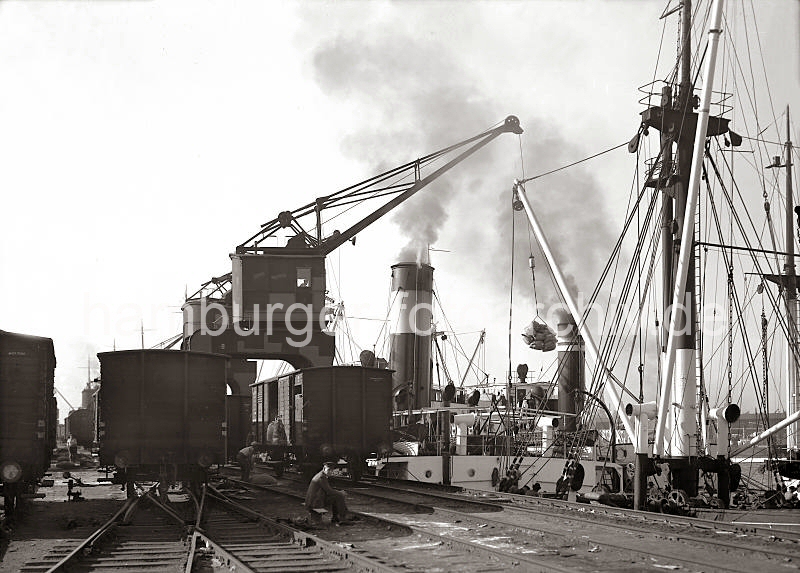 Historische Motive aus dem Hamburger Hafen, Freiladeverkehr am Holthusenkai der Norderelbe - Gterwaggons der Hafenbahn stehen auf den Gleisen - Portalkrane. 540_138 Ein Frachter liegt an der Kaimauer des Holthusenkais in der Norderelbe - ber einen Portalkran wird das Schiff mit Scken beladen. Auf den Gleisen der Bahnanlage stehen die Gterwaggons von denen die Fracht auf das Frachtschiff geladen wird.