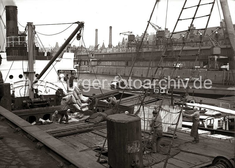 Historische Fotografien aus dem Hamburger Hafen / Bildarchiv der HLA. Stromumschlag im Hamburger Sandtorhafen am Kaiserkai - Hieve Scke am Ladebaum eines Frachters; ca. 1934.  541_158 Ein Frachters wird mit dem bordeigenen Ladebaum von lngsseits liegende Binnenschiffen beladen - aus dem Laderaum des einen Motorschiffs ist gerade eine Hieve Scke zum Frachter transportiert worden; Schauerleute nehmen sie in Empfang und verstauen die Scke im Frachtraum. Rechts werden gerade zwei Kisten an Deck des Frachtschiffs gebracht, die von einer weiteren lngsseits liegenden Schute gebracht wurde. Auf der gegenber liegenden Seite des Hafenbeckens vom Sandtorhafen der Sandtorkai mit den Lagerschuppen und Krananlagen - im Hintergrund die Backsteingiebel der Speicherstadt und die zwei Schornsteine vom Kesselhaus, das die Speicherstadt mit Strom versorgte und die Dampfenergie erzeugte, mit denen die hydraulischen Winden, Krne, Treppenaufzge und Hebebhnen in der Speicherstadt betrieben werden.