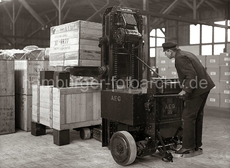 Historische Hamburgfotografien aus dem Hafen / Bilder von Hamburg als Geschenk zum Jubilaeum. Elektrohubwagen der Firma AEG - stapeln von Transportkisten im Lagerschuppen des Hafens. 547_186a Vorsichtig stapelt ein Arbeiter mit dem Elektrohubwagen zwei Holzkisten bereinander. Die Kisten stehen auf Blcken als Abstandshalter, damit die Gabel des AEG Elektrorwagens dazwischen gefahren werden kann.