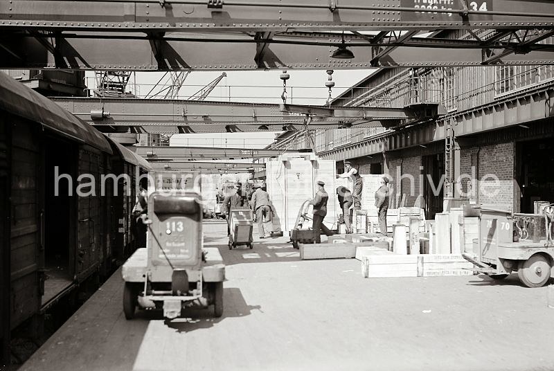 Historische Fotos von der Arbeit im Hafen. Fotoposter als Abschiedsgeschenk aus der Firma. Gueterzug an der Laderampe eines Hafenschuppens - Arbeiter mit Sackkarre + Elektrotransportkarren; ca. 1934.  550_392 Ein Gterzug mit geschlossenen Waggons steht an der Laderampe vor einem Hafenschuppen und wird entladen. Ein Arbeiter bringt eine grosse Holzkiste mit einer Sackkarre in das Lagerhaus, ein anderer fhrt mit seiner Karre zum Zug, um eine neue Ladung abzuholen. Mehrere Arbeiter lsen gerade die Seile an denen eine groe Transportkiste mit dem Kran auf der Rampe abgestellt worden ist. Im Vordergrund eine Elektrokarre, mit der kleinen Plattform auf dem der Fahrer stand und dem Rckenpolster, an dem er sich beim Fahren des Elektrotransportwagens absttzen konnte.