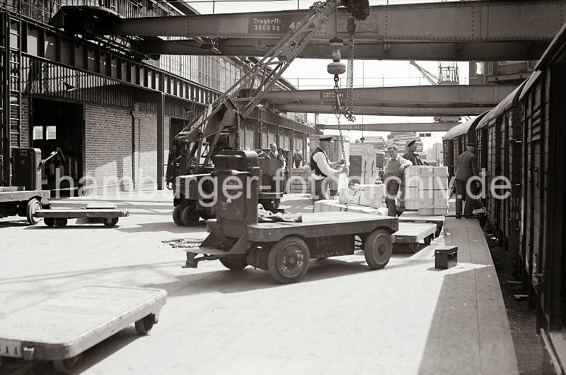 Historische Bilder von der Arbeit im Hafen / Fotos aus dem Hamburger Hafen als Firmengeschenk zum Abschied Mitarbeiter. Gueterwaggons an der Laderampe; ca. 1934. 551_393 Gterumschlag auf einer Laderampe des Hamburger Hafens. Transportkisten liegen auf einem Anhnger des Elektrokarrens - Hafenarbeiter stehen mit ihren Sackkarren vor dem Gterwaggon. Ein Arbeiter lst gerade das Ladegeschirr einer Hieve Kisten, die an Land gebracht wurde. Im Hintergrund sind Kisten und Baumstmme auf der Rampe zu erkennen.
