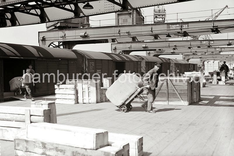 istorische Fotos aus der Arbeitswelt des Hafens. Hamburg Bilder als Dekoration im Buero. Entladung eines Gueterzugs - Transport von Ballen auf der Sackkarre, Holzkisten auf der Laderampe; ca. 1934.  552_393a Ein Gterzug steht an einer Laderampe des Hamburger Hafens und wird von Hafenarbeitern entladen. Zwei der Arbeiter ziehen ihre Sackkarre mit einem grossen Ballen ber die Rampe Richtung Lagerschuppen. Holzkisten in unterschiedlichen Grssen warten auf den Abtransport, im Hintergrund steht der Fahrer eines Elektrokarrens auf dem Fhrerstand seines Fahrzeugs.