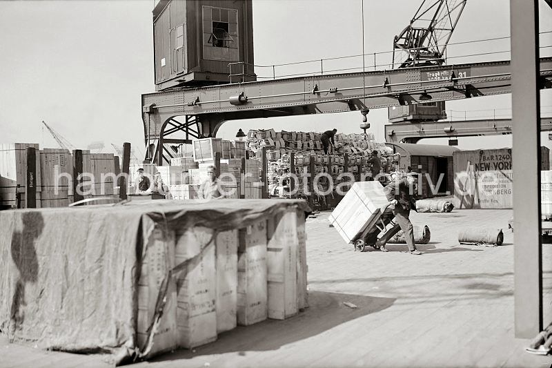 Alte Bilder aus der Geschichte des Hafen Hamburgs / Gterzug an der Laderampe im Hamburger Hafen - Hafenarbeiter mit Sackkarre + Kiste. Fotos von Hamburg als Weihnachtsgeschenk. 553_391a Ein mit Kisten und Sthlen hochbeladener Gterzug steht an einer Laderampe - der die Eisenbahnwaggons werden entladen; ein Arbeiter transportiert mit einer Sackkarre eine grosse Holzkiste zum Lagerschuppen. Im Vordergrund sind Kisten auf der Rampe gestapelt, die zum Schutz mit einer Plane abgedeckt sind.