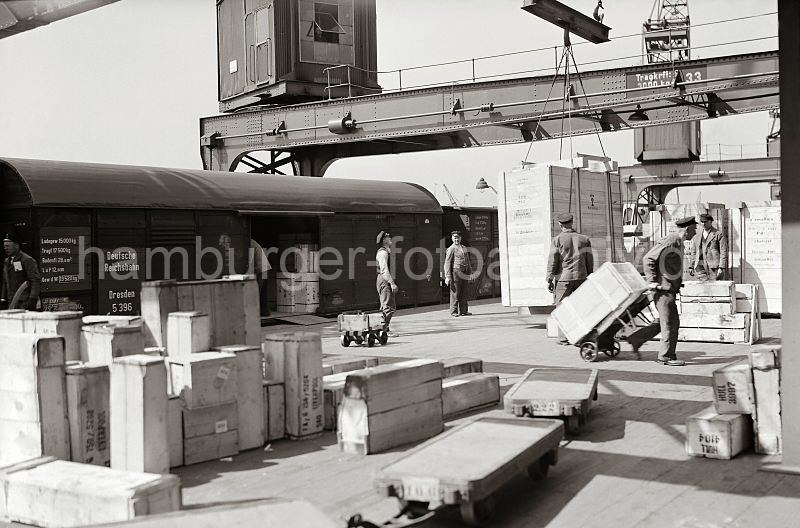 Historisches Bildmaterial vom Hafen Hamburgs - Holzkisten auf einer Laderampe im Hamburger Hafen - Hafenarbeiter beim Gterumschlag. Bilder als Weihnachtsgeschenk Hamburg.  554_392a Holzkisten in unterschiedlichen Grssen sind auf der Laderampe gestapelt - eine der Kisten wird mit der Handkarre in das Lagerhaus gebracht, eine weitere Transportkiste hngt am Haken des Halbportalkrans. Mehrere Hafenarbeiter sind an dem Ladevorgang beteiligt; im Vordergrund leere Anhnger von Elektrokarren.
