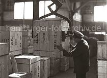 562_59 Eine Transportkiste hängt am Kistengreifer des Schuppenkrans; ein Lagerarbeiter dirigiert die Holzkiste an ihren Lagerplatz im Hafenschuppen.