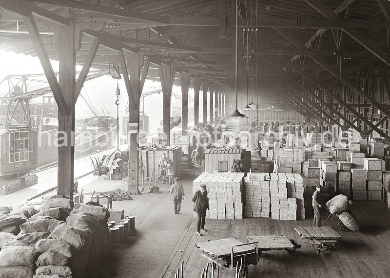 Historische Bilder von der Hafenarbeit in Hamburg; Warenumschlag im Kaischuppen 14, Dalmankai im Hamburger Grasbrookhaen; Hamburg-Bilder als Mitarbeiter-Geschenk zum Abschied. 566_0672a Die unterschiedlichsten Waren sind im Lagerschuppen 14 des Dalmankais im Grasbrookhafen gestapelt. An einem der Kaikrane, die von Laufschienen entlang der Kaimauer gefhrt werden hngt eine Ladung Kisten, die auf der Laderampe abgesetzt werden soll. Ein Hafenarbeiter wartet mit seiner Sackkarre darauf, die Holzkisten in den Lagerraum zu bringen.