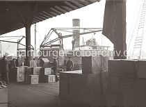 569_653 Hafenkrane entladen ein Frachtschiff - Hafenarbeiter stapeln die angelandeten Kisten von Hand in dem Lagerschuppen des Hamburger Hafens.  Auf den Kisten ist der Name der Lieferfirma Avreboo Rubber Co. Ltd. aufgedruckt.