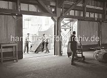 571_658 Hafenarbeiter transportieren Kisten mit der Sackkarre auf der Laderampe und im Lagerschuppen. Im Hintergrund liegt ein Frachtschiff am Kai.