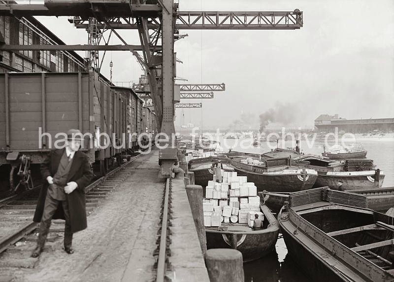 Bilder der Hafengeschichte Hamburgs - Fotos aus dem Archiv der HHLA; Kaianlage   am Afrikakai des S�dwesthafenens - G�terzug der Hafenbahn, Halbportalkrane. 579_645 Schuten liegen am Afrikakai des Hamburger S�dwesthafens - einige der Lastk�hne sind leer, w�hrend ein anderer mit Holzkisten hoch beladen ist. Unter den Halbportalkranen steht ein G�terzug; im Hintergrund dampft ein Schlepper in dem Hafenbecken. Der gegen�ber liegende Togokai  des ehemaligen Petroleumhafens ist noch nicht fertig gestellt .
