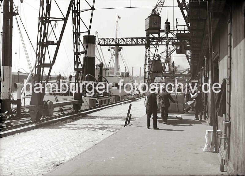 594_650 In Ein Frachtschiff mit hohem Schornstein liegt an einem Kai im Hamburger Hafen, die Ladung des Frachters wird gelscht; gerade wird eine Hieve mit Kisten von einem der Halportalkrane auf der Laderampe abgesetzt. Im Hintergrund transportieren Hafenarbeiter mit Sackkarren die Gter in den Lagerschuppen.  Alte Fotos aus dem Hamburger Hafen - Bildarchiv der HHLA. Die Ladung eines Frachters wird gelscht - Kisten auf der Laderampe.