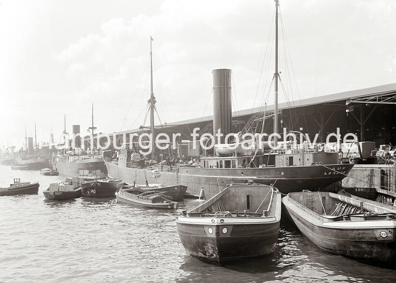 Alte Hafenbilder aus dem Fotoarchiv der Hamburger Hafen und Logistik AG (HHLA. Motorschiffe und Lastkhne am Dalmannkai des Grasbrookhafens; ca. 1932.  600_671 Die Ladung eines britischen Frachtschiffs wird am Dalmannkai des Hamburger Grasbrookhafens gelscht. ber schiffseigene Ladebume werden Scke in die lngsseits liegenden Schute verladen. Ein weiterer Lastkahn ist mit Kisten hoch beladen - ein Schlepper fhrt gerade heran, um eine der Schute weg zu schleppen. Auf dem Dalmankai stehen Pferdefuhrwerke und Lastkraftwagen und warten auf Ladung.