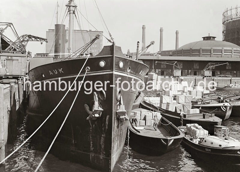 Hafenfotos von der Hansestadt Hamburg - Bildarchiv der HHLA - Frachtschiff am Dalmannkai - Schuten mit Ladung / Gaswerke Grasbrook; ca. 1936. 603_672 Der britische Frachter AUK liegt am Dalmannkai des Hamburger Grasbrookhafens. Hafenkrane lschen die Ladung des Frachtschiffs - auf der Wasserseite wird ber einen bordeigenen Ladebaum eine Hieve Kartons auf eine lngsseits liegende Schute geladen. Im Hintergrund rechts ein Ausschnitte des Gasometers vom Gaswerk Grasbrook, links das vom Hamburger Oberbaudirektor Fritz Schumacher entworfene Heringskhlhaus.