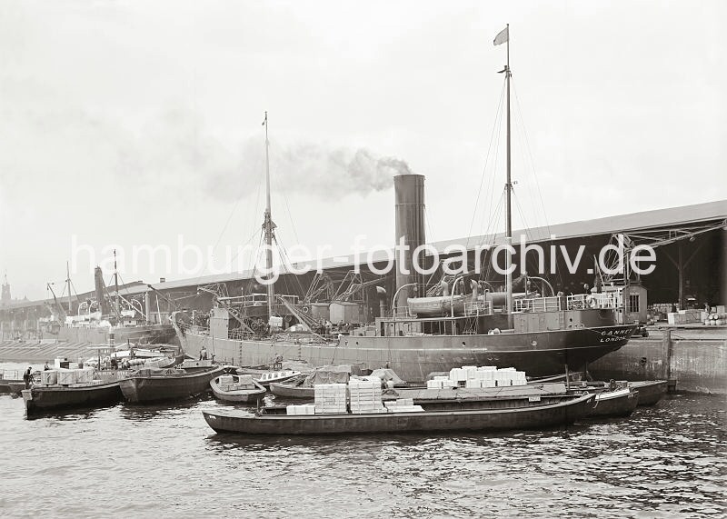 Hafenbilder von der Hansestadt Hamburg - Fotoarchiv der HHLA: Frachtschiff am Dalmannkai - Schuten mit Ladung / Gaswerke Grasbrook; ca. 1936. 604_597 Der Frachter GANNET mit Heimathafen London liegt im Grasbrookhafen am Dalmannkai und wird be- und entladen. Mehrere beladene Schuten haben am Schiff fest gemacht. An dem Bordkran des Frachters hngt eine Ladung Kisten. Ganz lks. im Hintergrund der Turm des Kaiserspeichers.