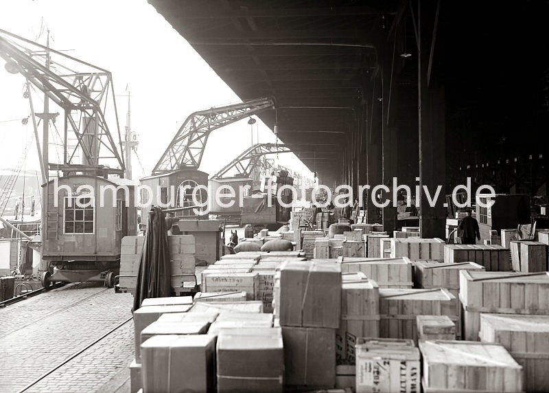 Historisches Bildmaterial aus der Hafenstadt Hamburg - Bildarchiv der HHLA. Kaischuppen 15 am Dalmannkai, Hafenkrane auf Schienen - Pferdefuhrwerk an der Laderampe. 606_669 Kaibetrieb am Dalmannkai im Grasbrookhafen - Schienenkrane lschen die Ladung eines Frachters, eine Hieve Kisten wird gerade auf der Laderampe des Kaischuppen 15 abgestellt. Die Lagerflche ist mit Kisten, Kartons und Scken vollgestellt. Ein Pferdfuhrwerk steht an der Rampe und im Hintergrund wartet ein Lastkraftwagen und ein Gterwaggon darauf, beladen zu werden.