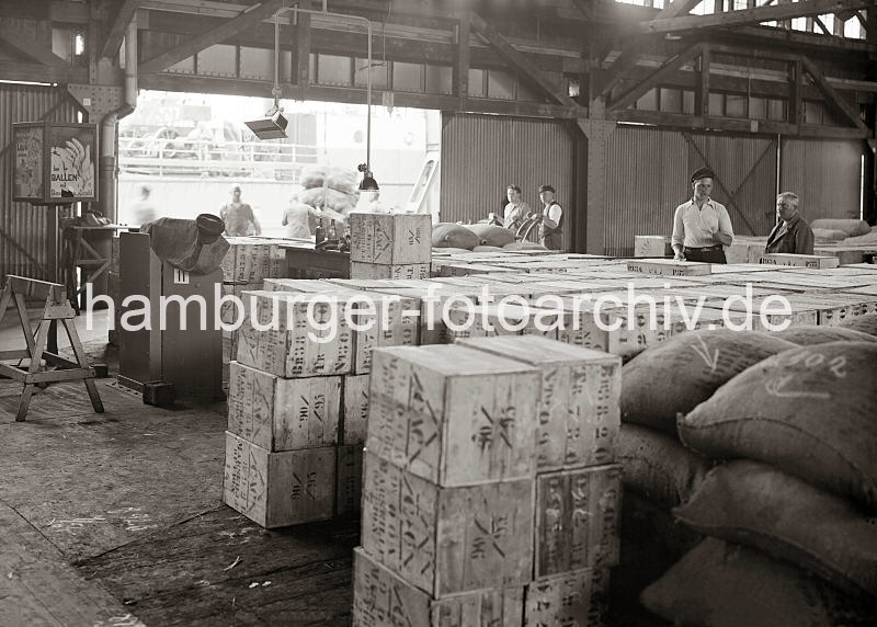 Altes Fotomaterial aus der Hafenstadt Hamburg - Bildarchiv der HHLA. Lagergut im Kaischuppen 15 am Dalmannkai, Holzkisten + Scke - Hafenarbeiter mit Sackkarren; ca.1936. 607_673 Blick in den Lagerschuppen 15 am Dalmannkai - Holzkisten und Scke sind in dem Lagerraum gestapelt - Hafenarbeiter schieben leere Sackkarren zur Laderampe. Links weist ein Schild auf Verletzungsgefahr hin, die durch Ballen mit Bandeisen und Draht entstehen knnen und dass nicht unter der schwebenden Last gestanden werden soll.