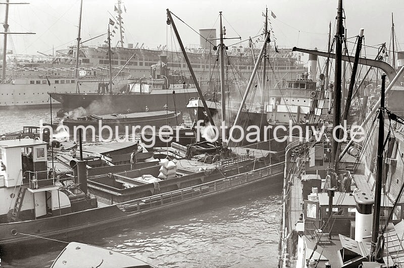 Bilder  von der Geschichte des Hamburger Hafens - Fotoarchiv der  HHLA. Beladung  eines Motorschiffs am Baumwall / Vorsetzen - NS-Passagierschiff Wilhelm Gustloff; ca.1939.  610_387 Ein Motorschiff wird vor den Kaianlagen an den Vorsetzen / Baumwall mit Scken beladen. Am bordeigenen Ladebaum hngt eine Ladung Scke, die von der lngsseits liegenden Schute geholt wurde. ber den Ausleger des Kaikrans wird der vordere Laderaum des Schiffs befrachtet. Im Hintergrund liegt das 1938 gebaute NS-Passagierschiff Wilhelm Gustloff am Ponton.