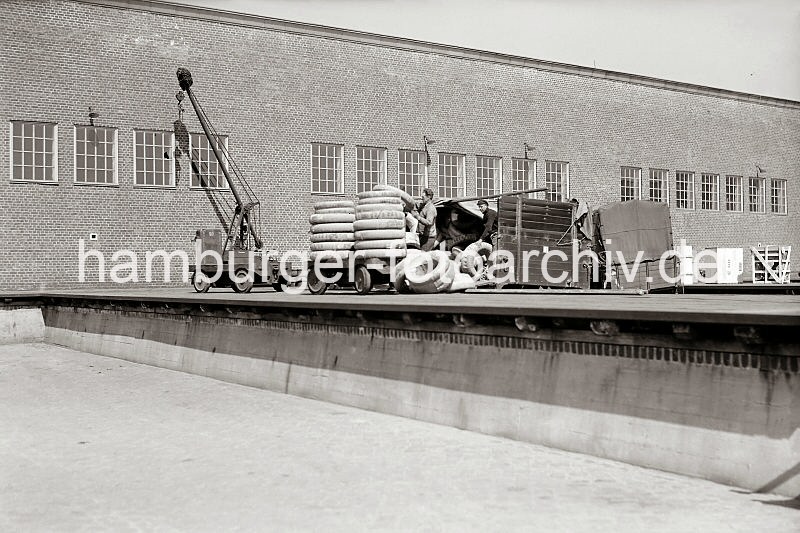 Foto-Dokumentation der Geschichte des Hamburger Hafens - Bildarchiv der HHLA - Verteilungsschuppen 57, Kamerunkai / Indiahafen - Entladen eines LKW mit Anhnger. 612_389 Auf der Laderampe des Verteilungsschuppens 57 am Kamerunkai des Indiahafens beladen Arbeiter den Anhnger eines Elektrokarrens mit Gummireifen - diese sind zum Schutz beim Transport in Papier gewickelt. Die Plane der Ladeflches des Lastkraftwagens ist zurckgeschlagen, einer der Arbeiter reicht dem Kollegen auf der Rampe die einzelnen Reifen. An der Wand steht ein fahrbarer kleiner Lagerkran.
