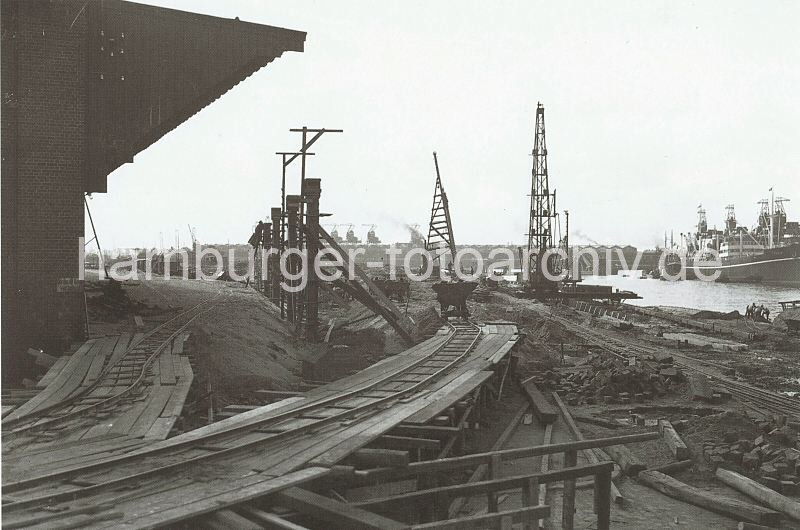 Historische Bilddokumente aus dem Fotoarchiv der Hamburger Hafen und Logistik AG HHLA. Baustelle am Togokai des Sdwesthafens - Erweiterung des Verteilungsschuppens 57; ca. 1934.  621_03502_58 Der alte Petroleumhafen wurde ab der 1920er Jahre zum Sdwesthafen umgebaut. Die Kaianlage des zuknftigen Togokais ist noch eine Baustelle. Provisorische Schienen sind auf Holzgersten verlegt - auf ihnen transportieren Bauloren Schutt und Material. Rechts liegen Frachtschiffe am Kamerunkai und im Hintergrund ist das tonnengewlbte Dach des Schuppens 59 am Windhukkai zu erkennen.