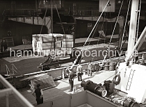 625_B_248a Blick über das Deck eines im Indiahafen liegenden Frachters. Eine Ladung Kisten schwebt über dem Laderaum des Frachtschiffs - im Hintergrund die Laderampe des Kaischuppens.