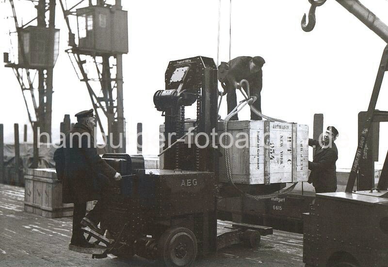 Historische Hamburg-Fotografien aus dem Bildarchiv der HHLA. Hubwagen der AEG mit Fahrer bei Verladung einer Holzkiste am Hafenkai; ca. 1932.  630_03502_30 Mit Hilfe eines Hubwagens wurde die groe Holzkiste aus dem Lagerschuppen auf den Hafenkai gefahren. Dort bereiten zwei Hafenarbeiter die Kiste fr die Verladung vor. Auf der Stirnseite der Kiste ist u.a. die Bezeichnung "Made in Germany" und der Bestimmungsort "New York" gedruckt.