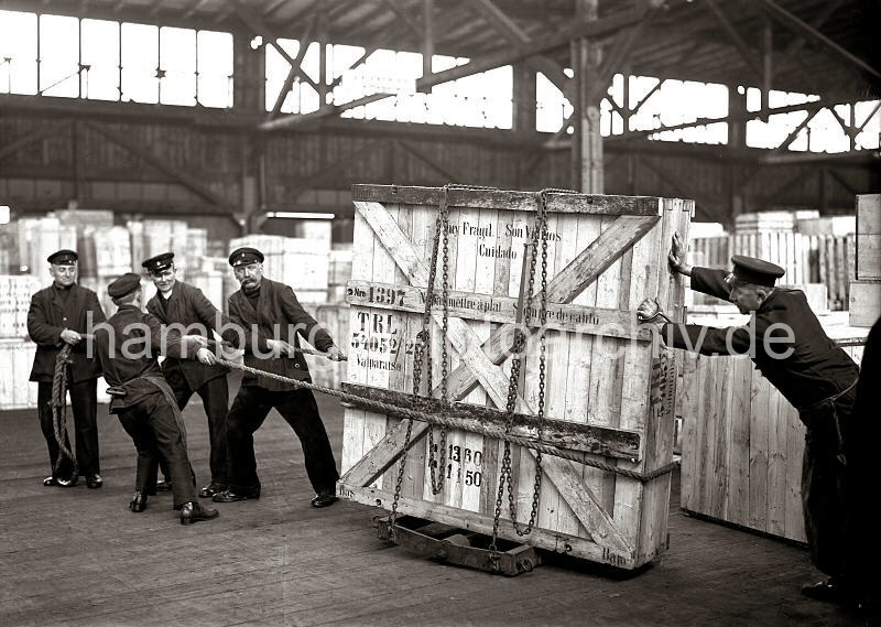 Historische  Arbeitsbilder aus dem Hafen Hamburgs - Bildarchiv der HHLA: Schwertransport einer Holzkiste per Schwerlastrollern - Hafenarbeiter im Hamburger Hafen; ca. 1932. 631_67 Fünf Mann arbeiten daran eine große Holzkiste mit Glasscheiben durch das Lager im Hamburger Hafen zu transportieren. Die Kiste steht auf Schwerlastrollen und ist mit einer Eisenkette an dem Gestell fixiert. Vier Männer ziehen die Ladung mit einem Seil, ein weiterer Lagerarbeit schiebt und hält die Balance der Kiste.
