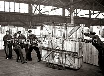 631_67 Fünf Mann arbeiten daran eine große Holzkiste mit Glasscheiben durch das Lager im Hamburger Hafen zu transportieren. Die Kiste steht auf Schwerlastrollen und ist mit einer Eisenkette an dem Gestell fixiert. Vier Männer ziehen die Ladung mit einem Seil, ein weiterer Lagerarbeit schiebt und hält die Balance der Kiste.