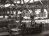 636_56 Mit einem Elektrolaufkran laden Arbeiter eine Partie Kupfer- barren auf den Anhänger eines Elektroschleppers. Das Hafenlager ist mit gestapelten Säcke und Baumwollballen gefüllt.